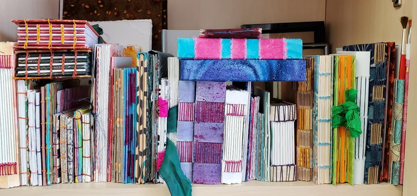A shelf filled with colorful, handmade books with visible bindings and vibrant cover designs. The scene conveys creativity and artisanal craftsmanship.